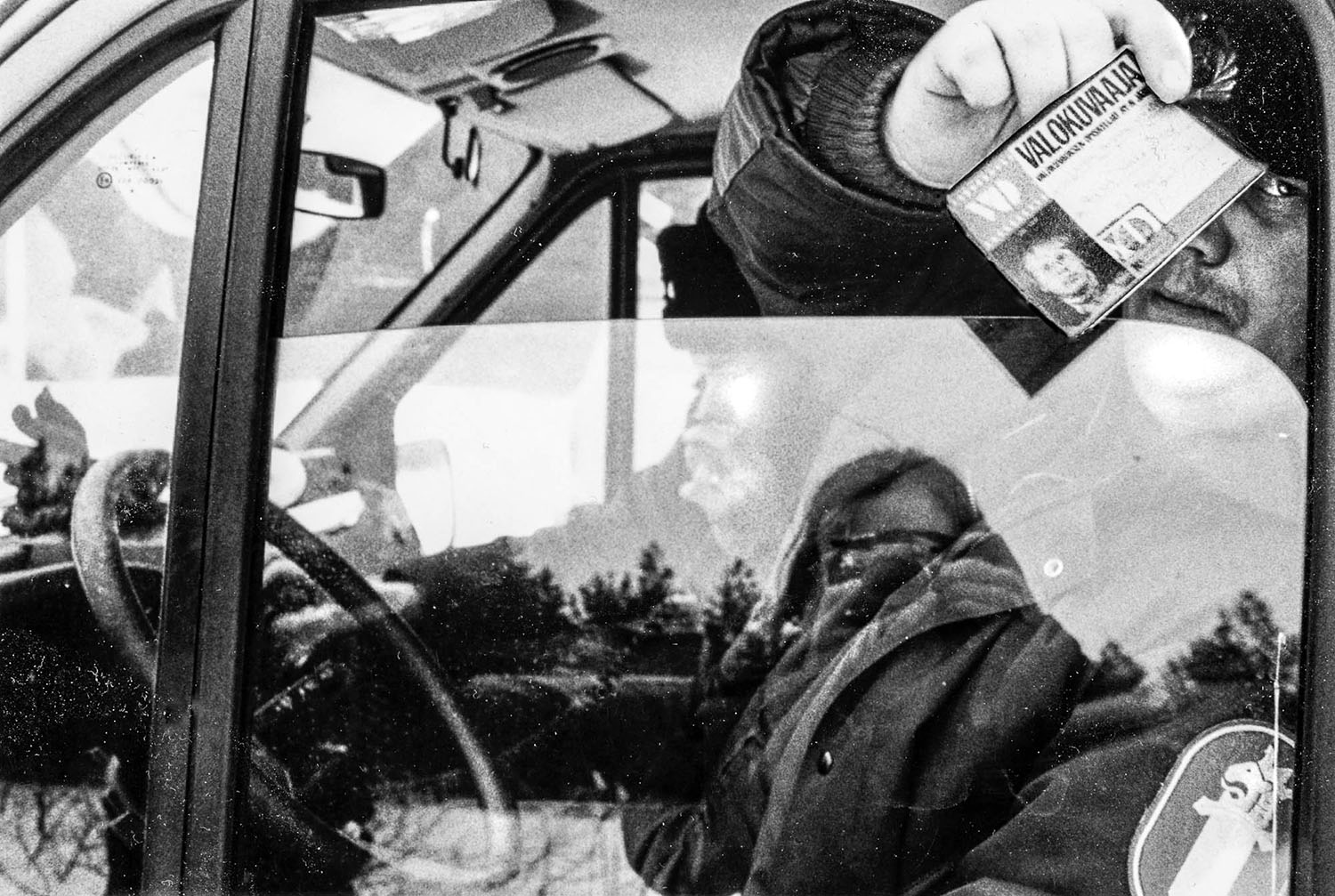 Lähikuvassa poliisiauto, jonka sisällä istuu kaksi poliisia. Pekka heijastuu kameran kanssa ikkunasta.